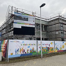 Bunte Farben auf einem Banner hängen vor dem Gerüst des Kita-Neubaus