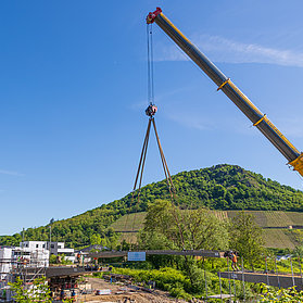Kran hebt Brückenteil als Teil des Einhubs vor Landschaft und blauem Himmel.