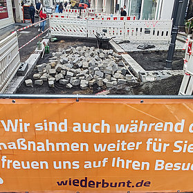 Offene Baustelle mit einem orangenen Banner davor: "Wir sind auch während der Baumaßnahmen weiter für Sie da und freuen uns auf Ihren Besuch."