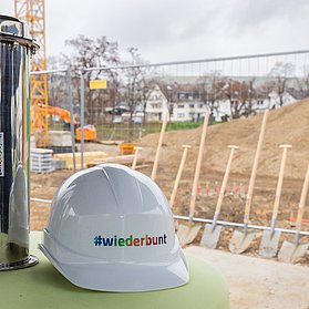 Zeitkapsel und #wiederbunt-Baustellenhelm vor der Baustelle der Feuerwehr Ahrweiler.