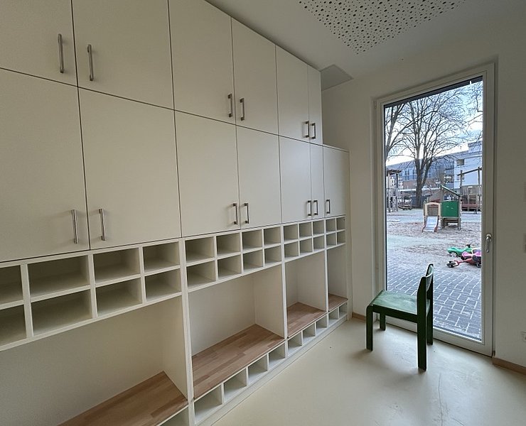 Ein Stuhl steht neben leeren Schränken und Regalen. Eine Glastür führt nach draußen zum Spielbereich.