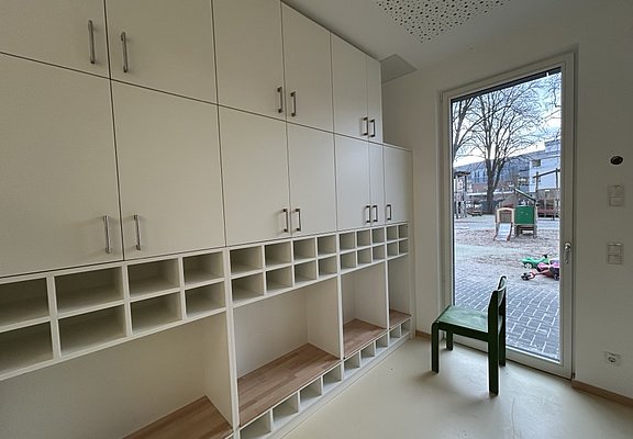 Ein Stuhl steht neben leeren Schränken und Regalen. Eine Glastür führt nach draußen zum Spielbereich.