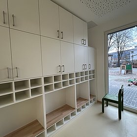 Ein Stuhl steht neben leeren Schränken und Regalen. Eine Glastür führt nach draußen zum Spielbereich.