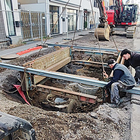 Zwei Arbeiter an schweren Metallteilen über einer offenen Baugrube.