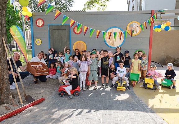 Die Kinder der Kita Arche Noah winken in die Kamera. Es ist mit Wimpeln geschmückt und die Kinder haben Spielzeugbagger und Bobbycars.