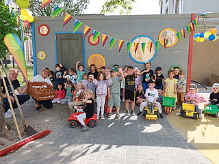 Die Kinder der Kita Arche Noah winken in die Kamera. Es ist mit Wimpeln geschmückt und die Kinder haben Spielzeugbagger und Bobbycars.