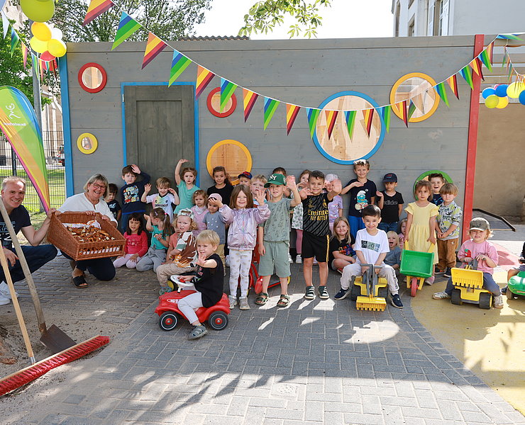 Die Kinder der Kita Arche Noah winken in die Kamera. Es ist mit Wimpeln geschmückt und die Kinder haben Spielzeugbagger und Bobbycars.