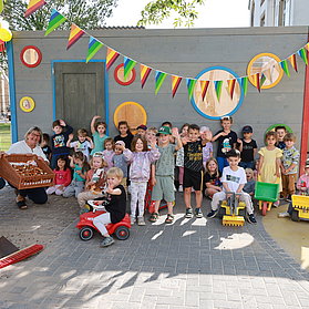 Die Kinder der Kita Arche Noah winken in die Kamera. Es ist mit Wimpeln geschmückt und die Kinder haben Spielzeugbagger und Bobbycars.