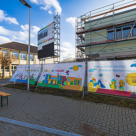 Bunte Farben auf einem Banner hängen vor dem Gerüst des Kita-Neubaus