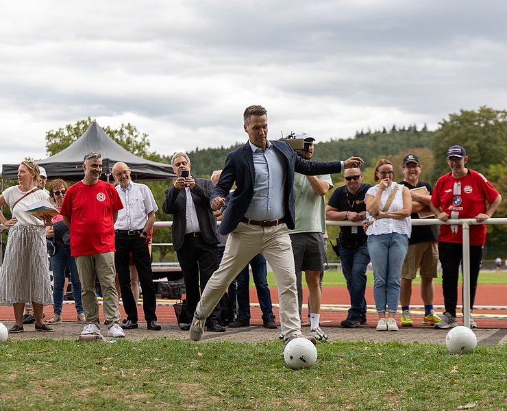 Der Bürgermeister schießt einen Fußball und wird dabei von Gästen beobachtet.