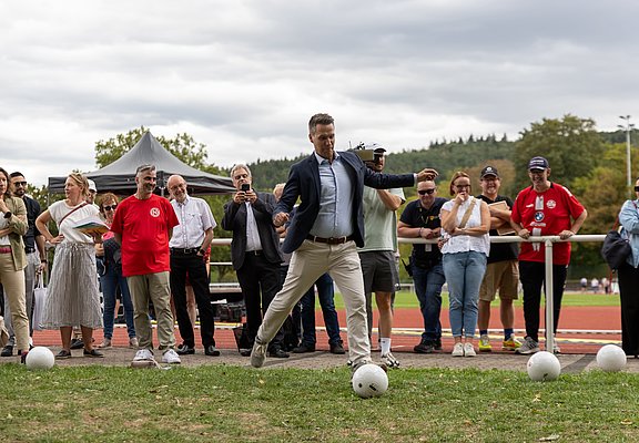 Der Bürgermeister schießt einen Fußball und wird dabei von Gästen beobachtet.